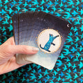 A hand holds a fanned deck of oracle cards featuring a night sky design with a blue cat silhouette on a moonlit background. The cards are shown against a dark patterned backdrop with teal bird silhouettes.