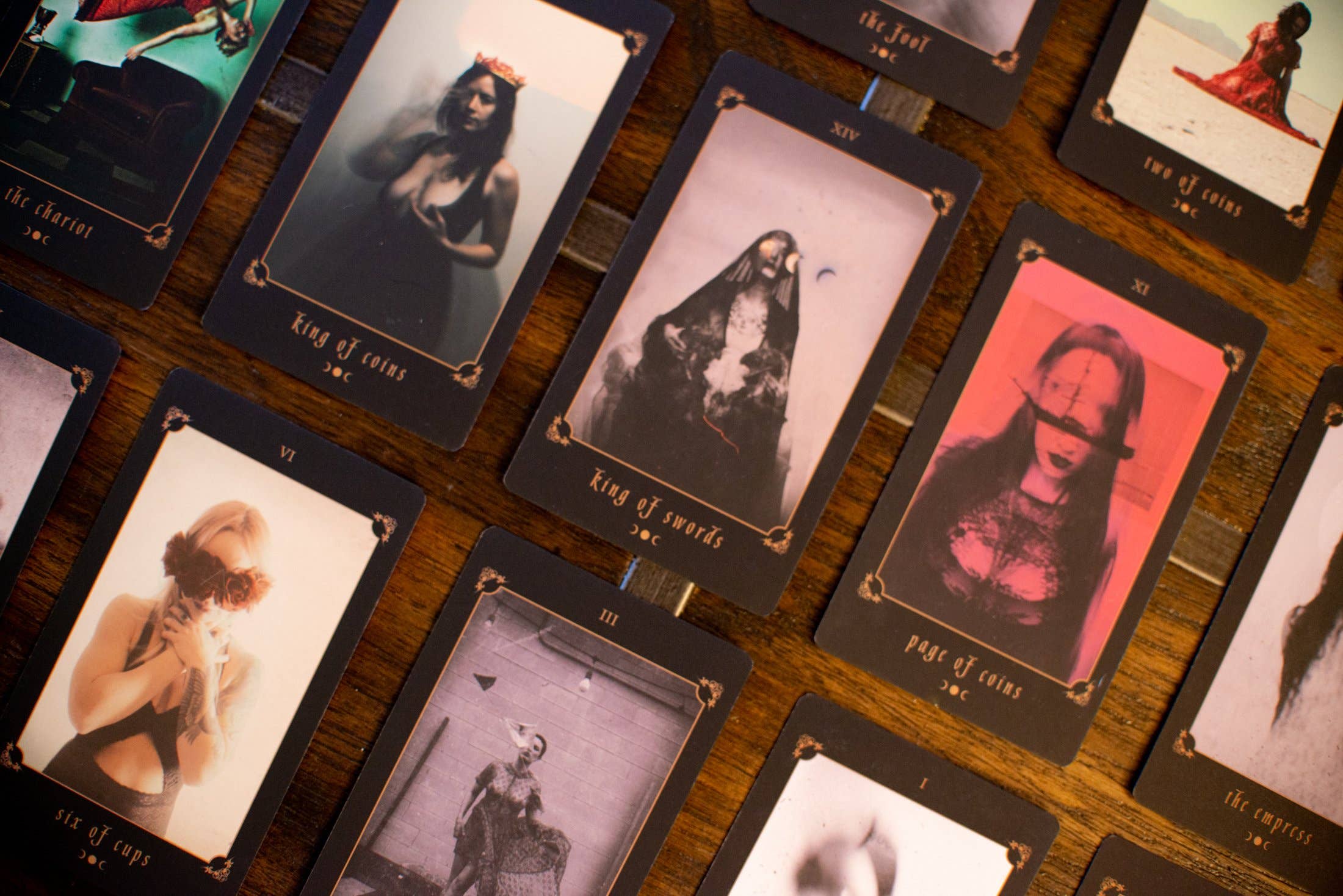 A close-up overhead view of tarot cards arranged on a wooden surface. The cards feature dark borders and stylized, moody portraits in muted and saturated tones, including pink, purple, black, and sepia imagery.