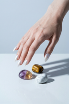 Hand with white nail polish touching three colorful crystals on a light blue background