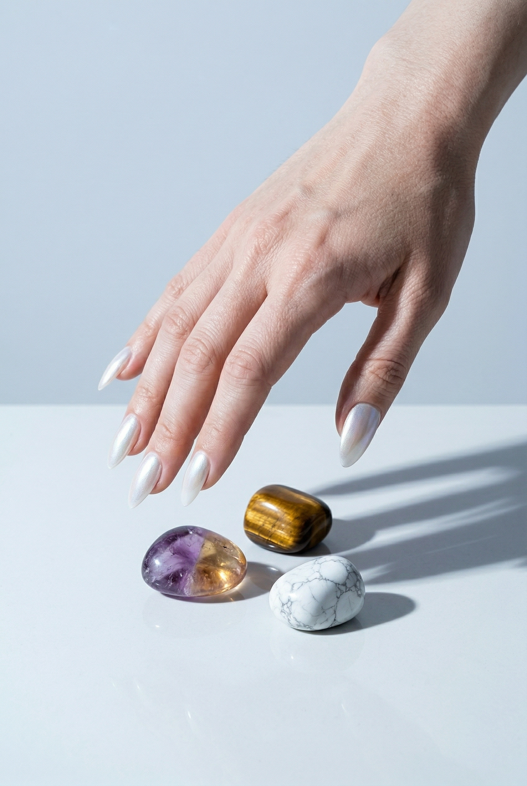 Hand with white nail polish touching three colorful crystals on a light blue background