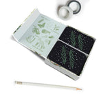 Set of black coasters with green designs in a box, accompanied by a pencil and tape on a white background.