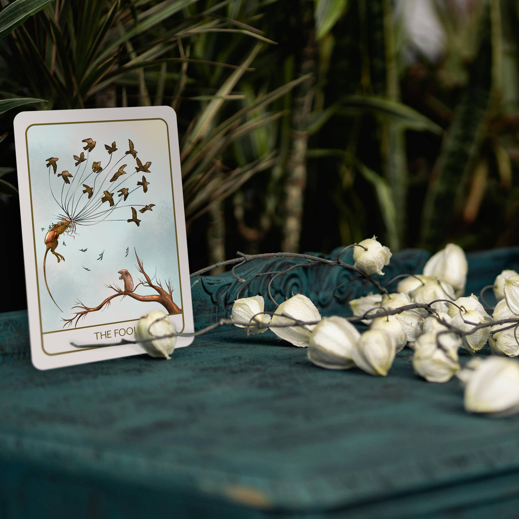 A tarot card titled 'THE FOOL' is displayed on a dark teal surface, surrounded by white dried flowers and green foliage in the background.