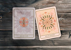 Two illustrated tarot-style cards, labeled Ace of Cups and Ace of Swords, placed on a rustic dark wood surface. The left card is pale gray with a peach and blue floral design, and the right card is pale pink with a peach, green, and blue radial floral design.