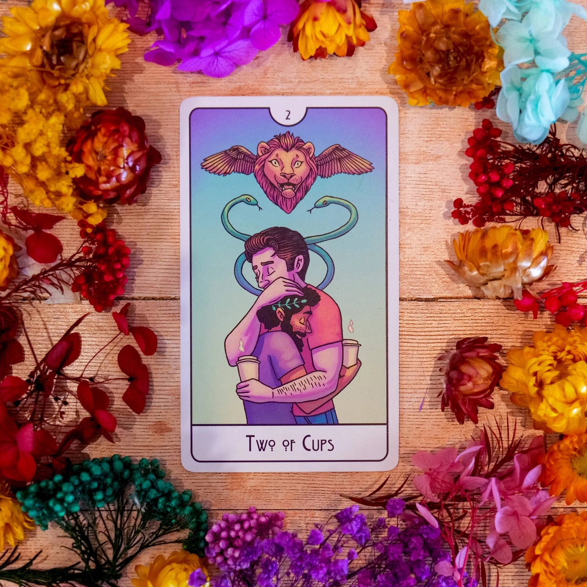 Two of Cups tarot card surrounded by colorful flowers on a wooden surface