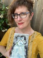 A woman with short brown hair and glasses holds up a watercolor painting of a fluffy cat. She is wearing a mustard yellow embroidered top and standing in front of a wooden fence with green plants on the left.