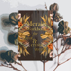 A rectangular workbook titled 'Merak Workbook for arot & crystal work' centered on a light gray background with botanical illustrations. The cover is dark brown with gold lettering and decorative gold and orange floral wreath graphics.