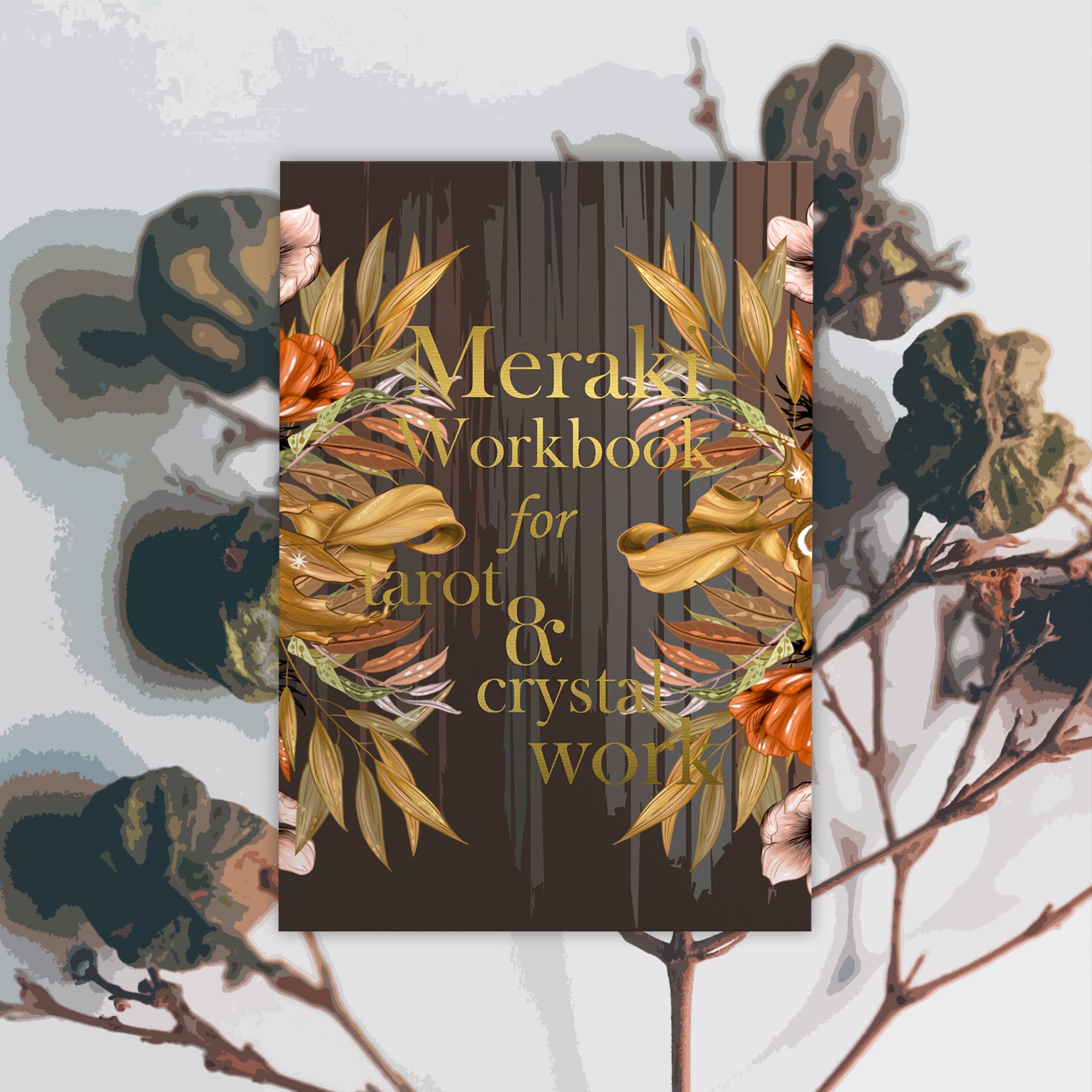 A rectangular workbook titled 'Merak Workbook for arot & crystal work' centered on a light gray background with botanical illustrations. The cover is dark brown with gold lettering and decorative gold and orange floral wreath graphics.