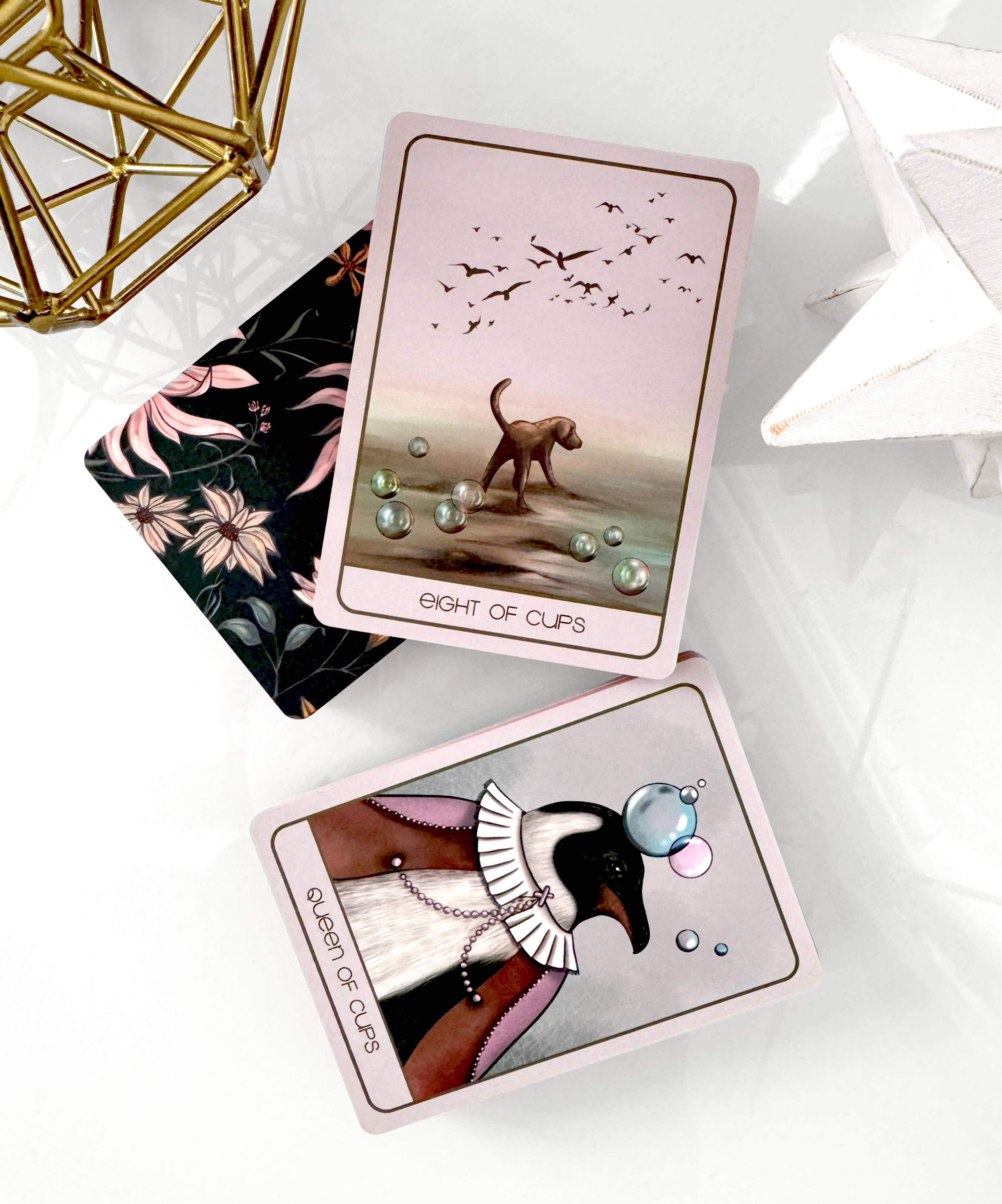 A styled flat-lay of a tarot card deck showing the 'Eight of Cups' and 'Six of Cups' cards on a glossy white surface, accented by a gold geometric object and a white star ornament.