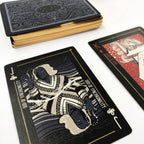 A deck of illustrated playing cards with a black-backed design and gold edges, shown with one face-up card displaying a stylized figure and the text 'GOD OF SYMMETRICALITY' and 'JACK OF WISDOM', plus part of another card with a red illustrated face.