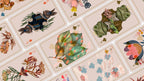 A close-up of illustrated playing cards arranged diagonally, showing whimsical nature-themed artwork including a rabbit, green frog, leaves, flowers, and birds on a pale pink card background.