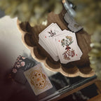 A styled flat-lay of a deck of illustrated playing cards on a scalloped wooden tray, with one card face showing a rabbit and another showing botanical roots, alongside a decorative card on a glass surface.