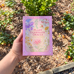 A hand holds a pink box of 'Plant Guardians Oracle' cards outdoors against a background of mulch and green plants. The box features colorful botanical illustrations and the brand 'Sow the Magic Co.
