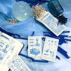 Blue-themed stationery items including cards, pencils, and a crystal ball on a blue fabric background.