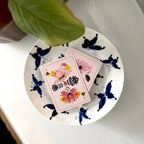 Two decorative playing cards with pink backgrounds and floral illustrations rest on a white ceramic plate decorated with blue butterflies. A green plant leaf is visible at the top edge of the frame.