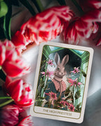 A tarot card titled 'THE HIGH PRIESTESS' featuring an illustrated rabbit in a green and pink Victorian-style scene, styled on a white surface with red tulips in the foreground.