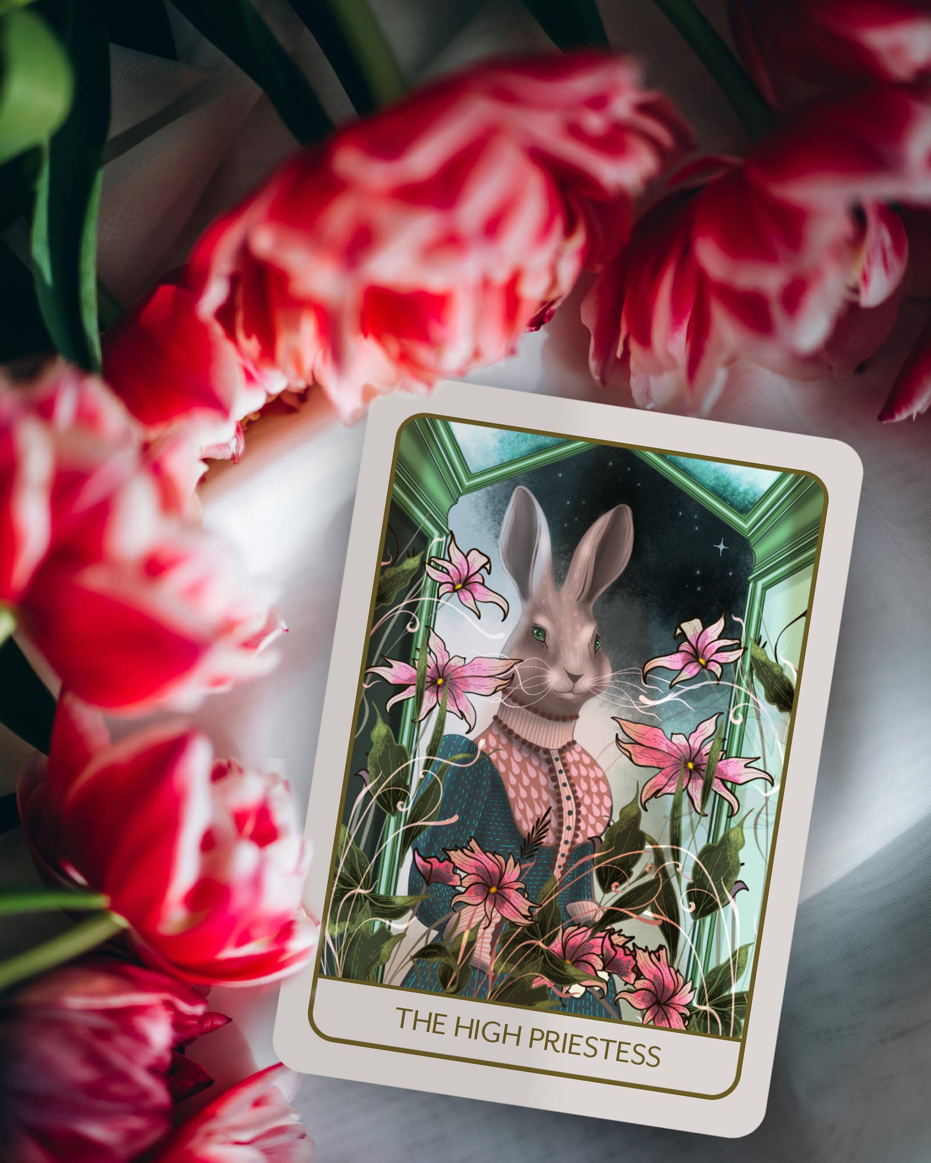 A tarot card titled 'THE HIGH PRIESTESS' featuring an illustrated rabbit in a green and pink Victorian-style scene, styled on a white surface with red tulips in the foreground.