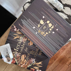 A close-up of a decorative book or workbook titled 'Meraki Workbook for growth & crystal work' with a dark brown cover featuring gold floral artwork and text. Another matching book is partially visible underneath, with crystals and a patterned rug in the background.