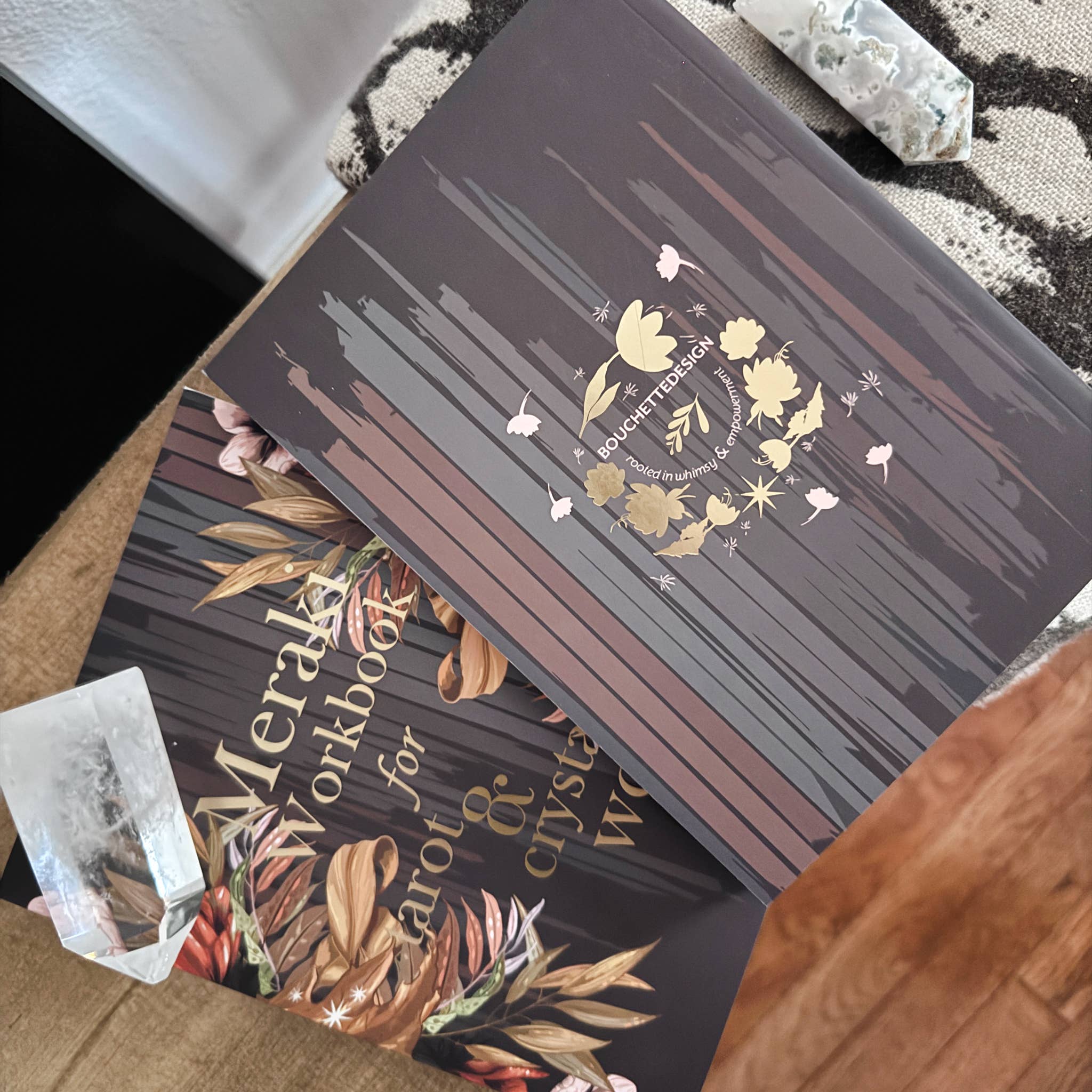 A close-up of a decorative book or workbook titled 'Meraki Workbook for growth & crystal work' with a dark brown cover featuring gold floral artwork and text. Another matching book is partially visible underneath, with crystals and a patterned rug in the background.