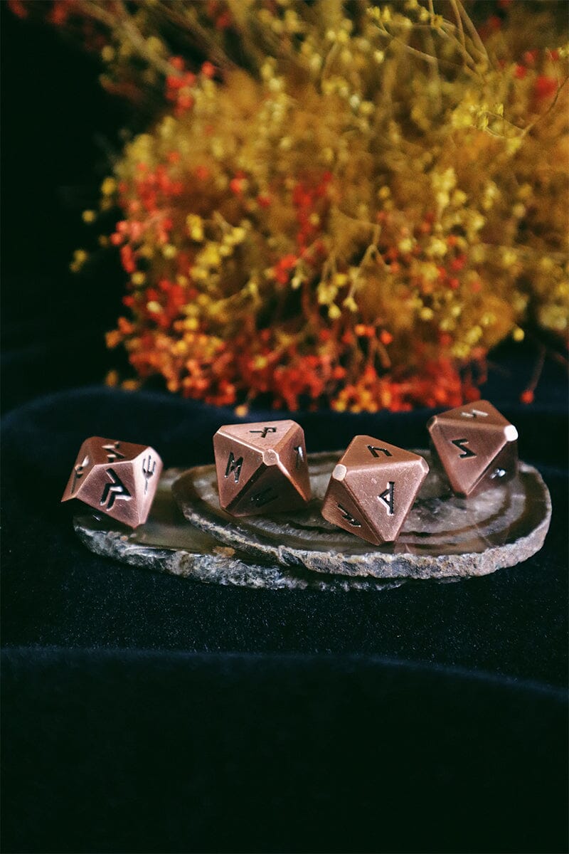 Set of five copper-colored polyhedral dice with symbols on a dark surface with a blurred natural background.