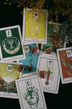 A styled flat-lay of multiple tarot or oracle cards spread on a dark surface, with a small potted plant and several pink dice. The cards feature illustrated symbols and text including 'ELUSIVE', 'INSPIRED', and 'SUGAR M'.