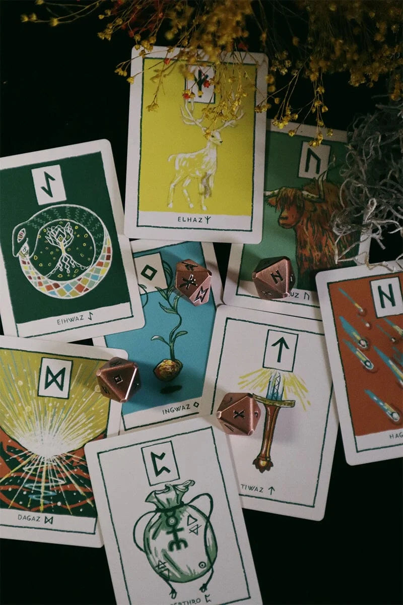 A styled flat-lay of multiple tarot or oracle cards spread on a dark surface, with a small potted plant and several pink dice. The cards feature illustrated symbols and text including 'ELUSIVE', 'INSPIRED', and 'SUGAR M'.