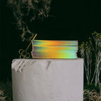 A stack of iridescent rainbow-colored cards or pages sits on a white pedestal, with a gold key resting against the left side. The background is very dark with dried plant elements.