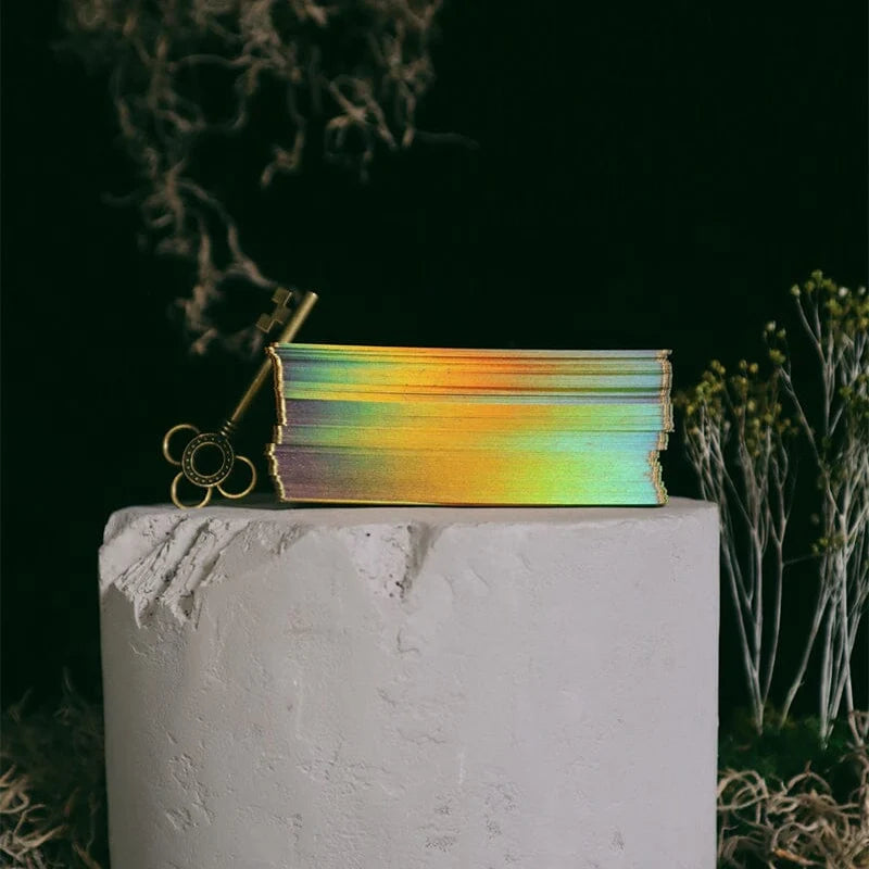 A stack of iridescent rainbow-colored cards or pages sits on a white pedestal, with a gold key resting against the left side. The background is very dark with dried plant elements.