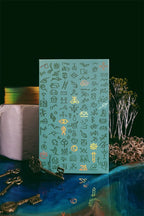 A rectangular teal card or plaque covered with small gold symbols and illustrations, standing upright on a glossy blue surface with decorative props including keys, a wrapped box, dried branches, and a dark green background.