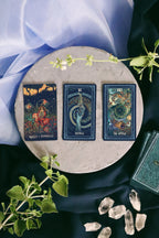 Three tarot cards on a round stone surface with crystals and greenery, on a dark fabric background.