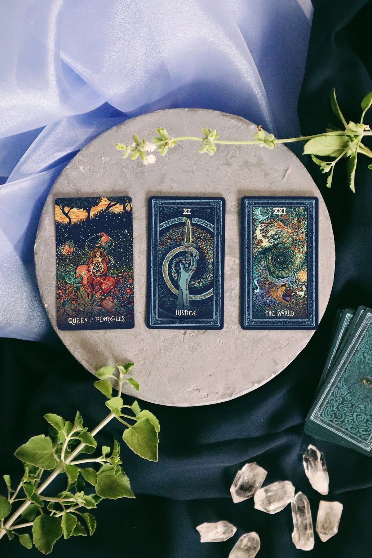 Three tarot cards on a round stone surface with crystals and greenery, on a dark fabric background.