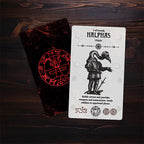 Card with a black and white illustration of a character holding a sword, placed on a wooden surface next to a black pouch with red symbols.